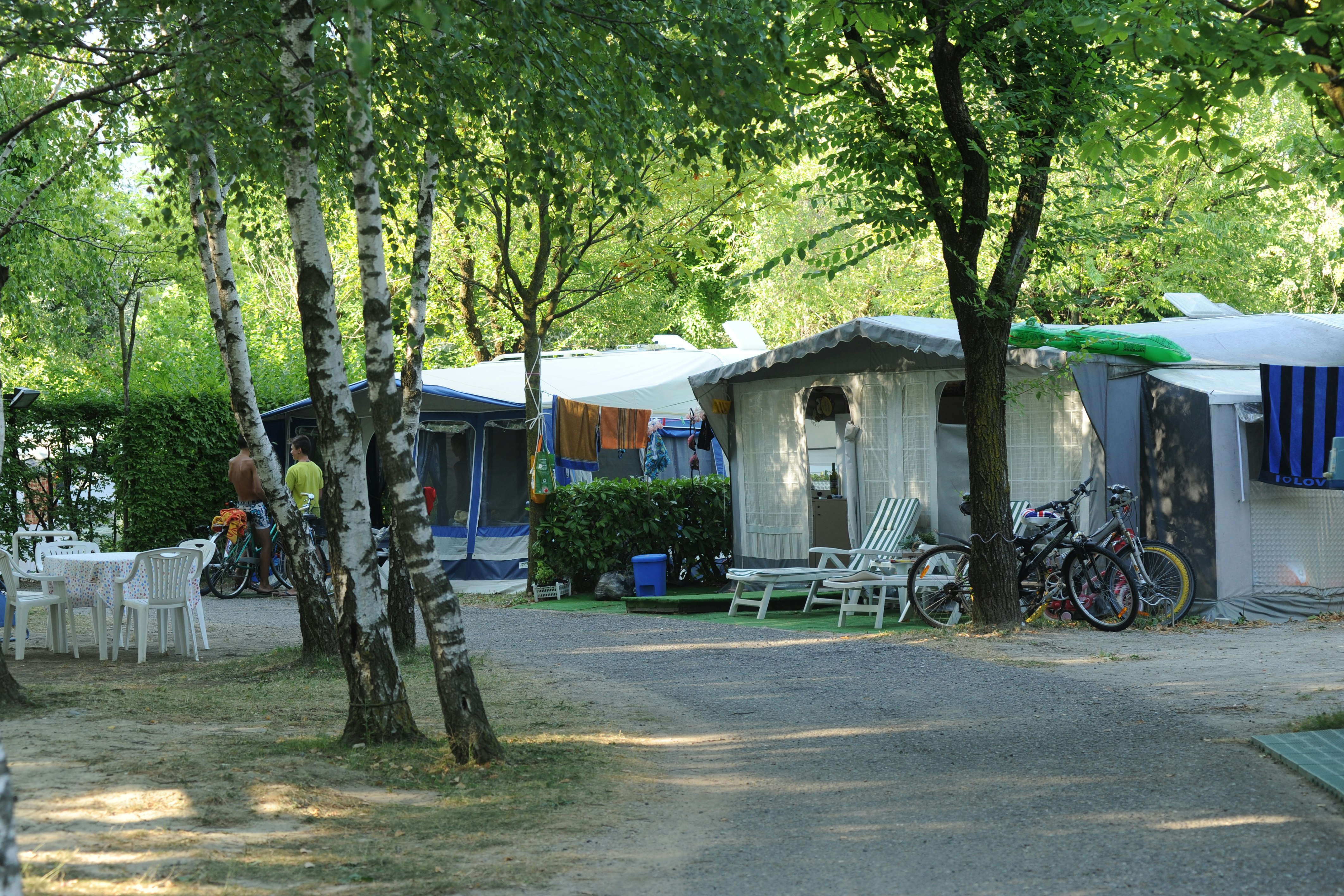 Campingplatz im Grünen: Zelte und Wohnwagen, weiße Sitzgarnitur am Weg, Fahrräder neben einem Wohnwagen unter Bäumen.