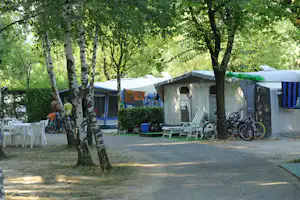 Campingplatz im Grünen: Zelte und Wohnwagen, weiße Sitzgarnitur am Weg, Fahrräder neben einem Wohnwagen unter Bäumen.