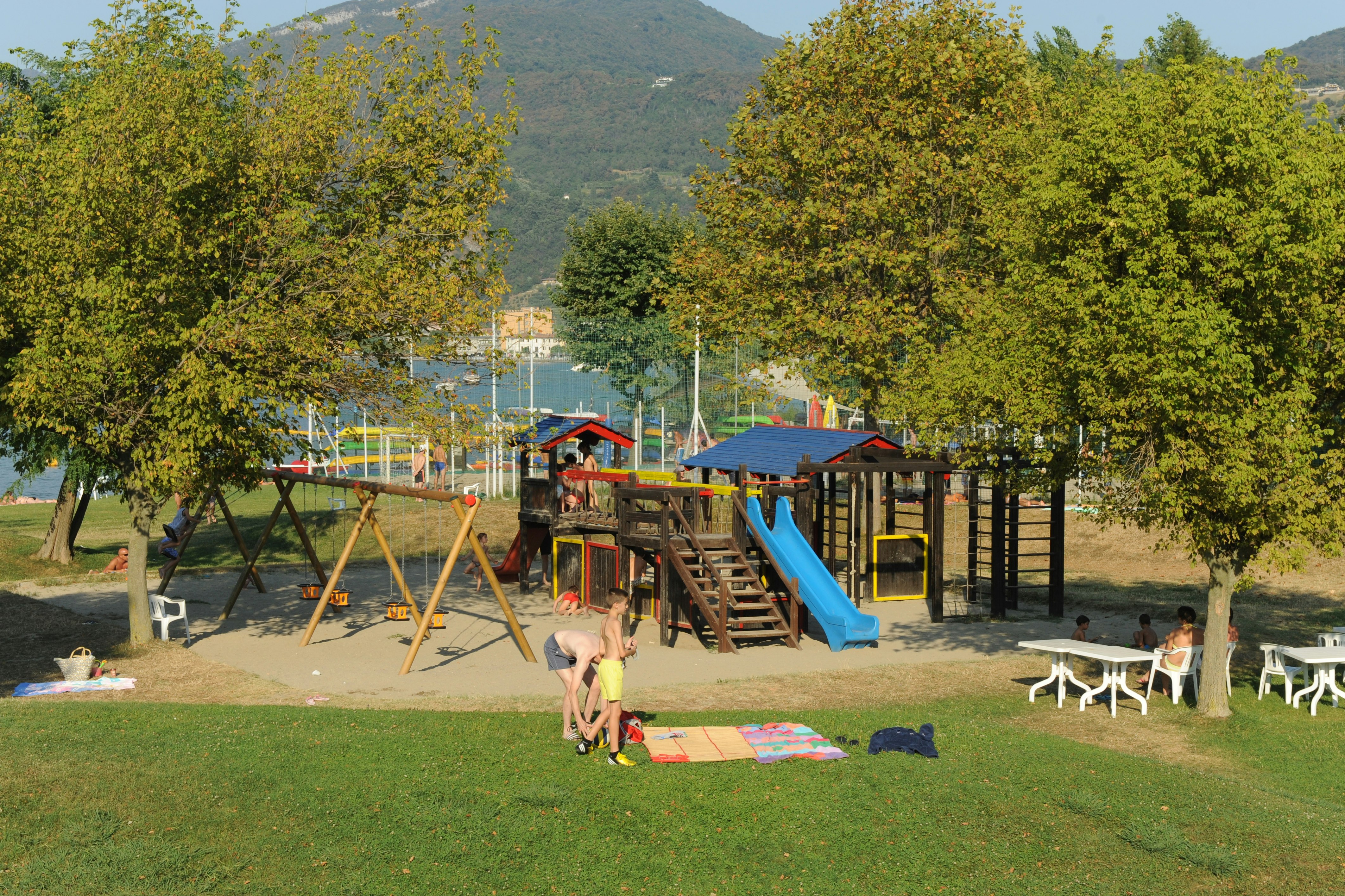Spielplatz auf dem Campingplatz mit Rutsche, Klettergerüst und Schaukel, umgeben von Bäumen und Blick auf die Berge.