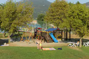 Spielplatz auf dem Campingplatz mit Rutsche, Klettergerüst und Schaukel, umgeben von Bäumen und Blick auf die Berge.