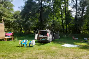 Stellplatz im Grünen mit Wohnwagen, geöffnetem Fahrzeug, Gartenmöbeln und Liegestühlen auf dem Rasen, umgeben von Bäumen.