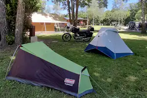 Zelte auf grünem Rasen im Campingbereich, umgeben von Bäumen; im Hintergrund ein Motorrad und ein weiteres Zelt.