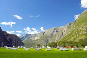 Campingplatz mit Wohnmobilen auf grünem Rasen, im Hintergrund Berge und ein See zwischen Felswänden.