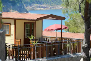Bungalow mit Holzterrasse, rotem Sonnenschirm und Blick auf den See, im Hintergrund Berge und Bäume.