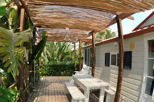 Terrasse eines Wohnwagens mit Holzdach, weißen Gartenmöbeln und Blick ins Grüne mit Palmen und Hecken.