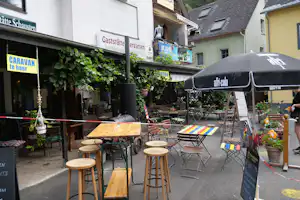 Terrasse mit Tischen und Hockern vor einer Gaststätte, Markisen und Häusern in einer kleinen Gasse, Pflanzen am Eingang.