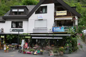 Pension und Gasthaus an einem Campingplatz, mit Schildern, Terrasse und Sitzgelegenheiten vor dem Gebäude.