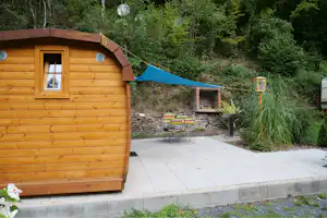 Holzhütte mit Fenster und Terrasse aus Steinplatten, im Hintergrund Bäume und ein Grill am Rand des Weges.