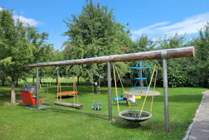Spielplatz am Campingplatz: Schaukelgestell mit mehreren Schaukeln, Trampolin und Rasen zwischen Bäumen.
