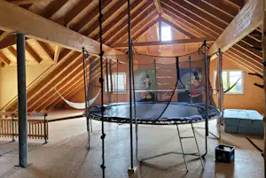 Trampolin mit Schutznetz in einem Holzhaus, Sicht auf Dachbalken, Fenster und Sitzbank im Innenraum.