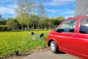Roter Camper am Stellplatz mit zwei Gartenstühlen auf grünem Rasen, Birkenreihe und blauem Himmel im Hintergrund.