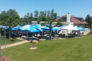 Terrasse mit blauen Sonnenschirmen, Tischen und Stühlen, Rasenfläche und Bäumen vor einem Gebäude mit rotem Dach.