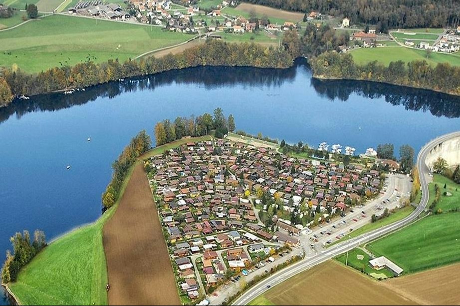 Luftaufnahme eines Campingplatzes am See mit vielen Stellplätzen, Wegen und Häusern am Ufer.