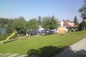Spielplatz mit Rutsche und Schaukeln auf grünem Rasen, im Hintergrund Gebäude und weitere Stellplätze am Campingplatz.