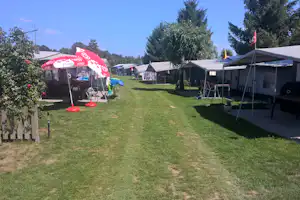Campingplatz mit Rasenweg, Zelt und Wohnwagen-Stellplätzen, daneben Bäume und ein Sonnenschirm mit buntem Muster.