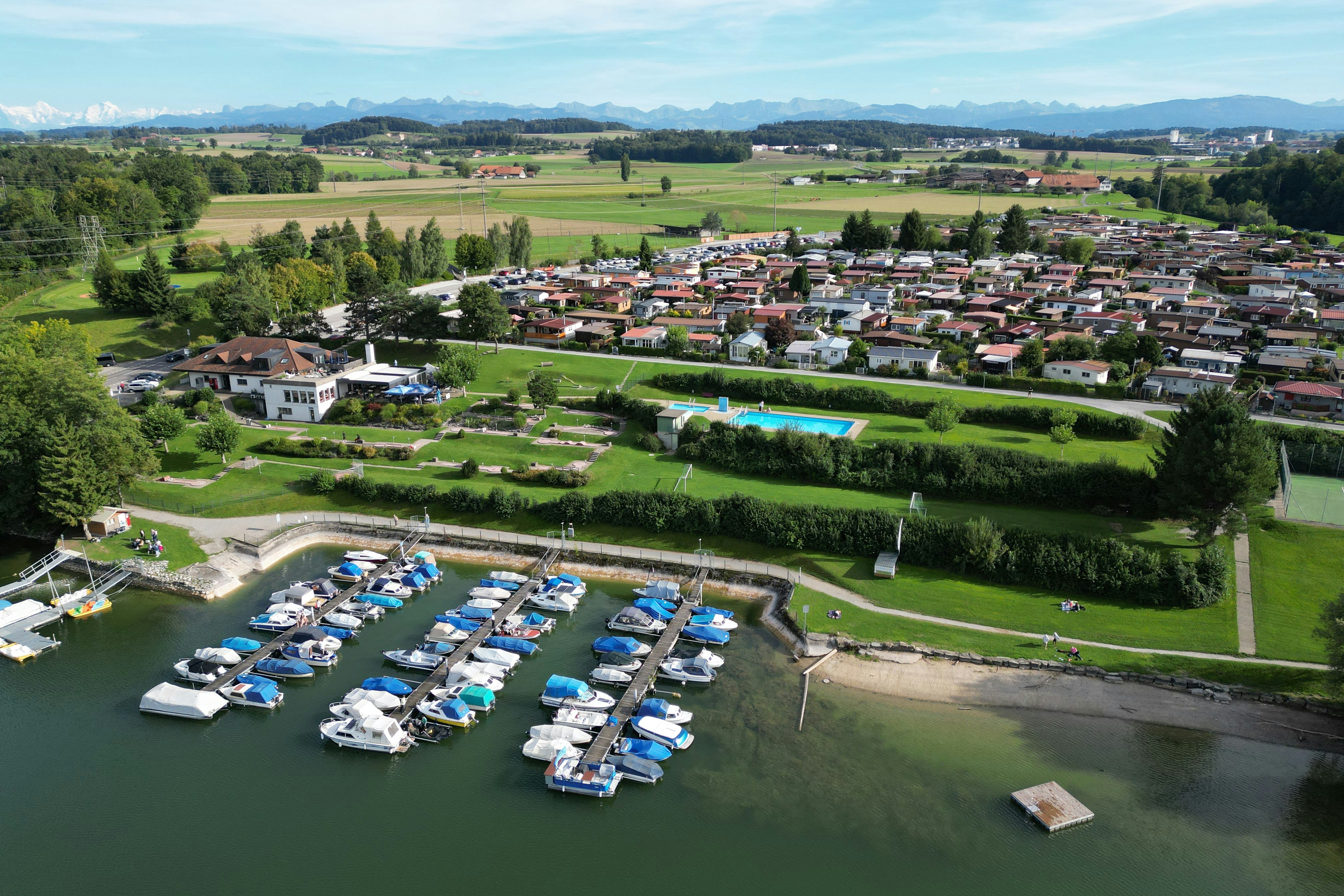 Luftaufnahme eines Campingplatzes am See mit Booten, Stellplätzen und Wohnwagen, umgeben von grünen Wiesen und Bäumen.