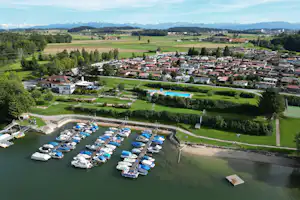 Luftaufnahme eines Campingplatzes am See mit Booten, Stellplätzen und Wohnwagen, umgeben von grünen Wiesen und Bäumen.
