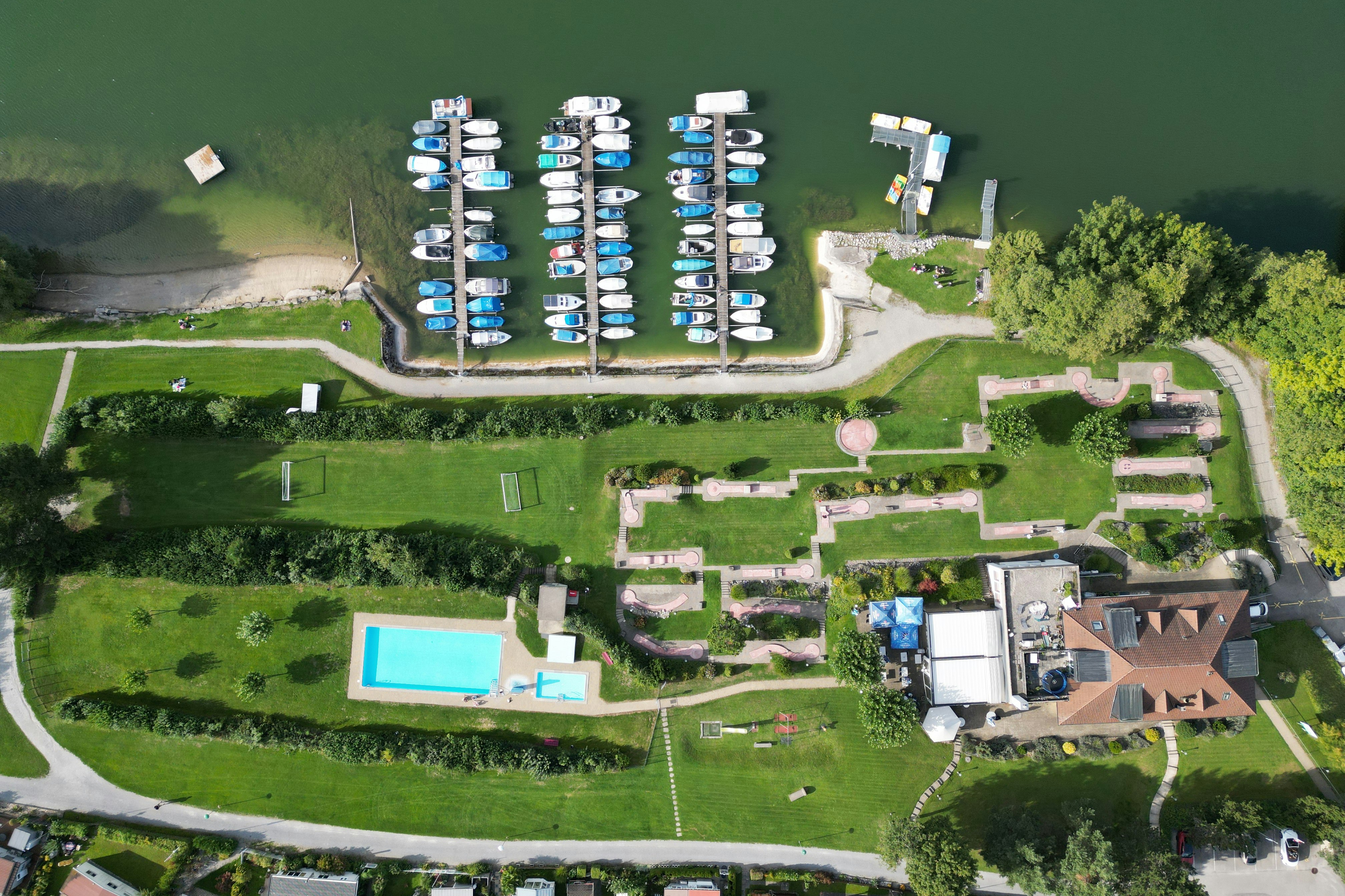 Luftaufnahme eines Campingplatzes mit mehreren Stellplätzen, Wohnmobilen und einem blauen Pool im grünen Gelände.