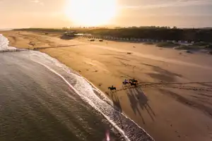 Weitblick auf einen Sandstrand mit Wellen, Pferden am Ufer und einem Campingbereich mit Zelten und Bungalows im Hintergrund.