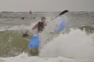 Zwei Surfer im Meer mit blauen Brettern, Wellen brechen am Strand, im Hintergrund weitere Personen im Wasser.
