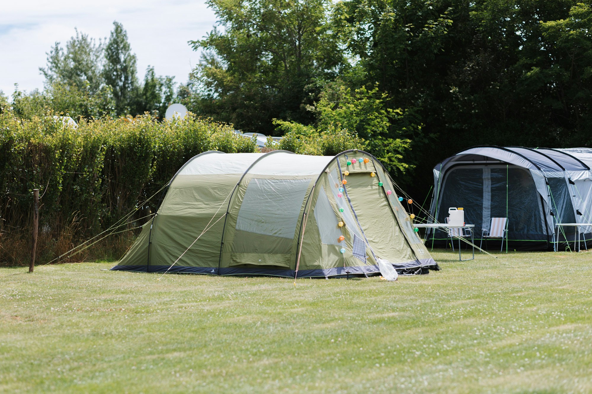 Zelte und ein Wohnwagen auf einer grünen Wiese im Campingbereich, umgeben von Büschen und Bäumen.