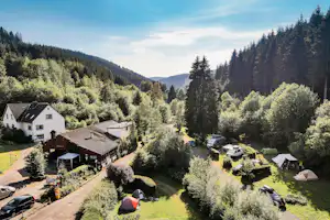 Campingplatz mit Zelten und Wohnwagen auf einer Wiese, umgeben von Bäumen und einem weißen Haus im Tal.