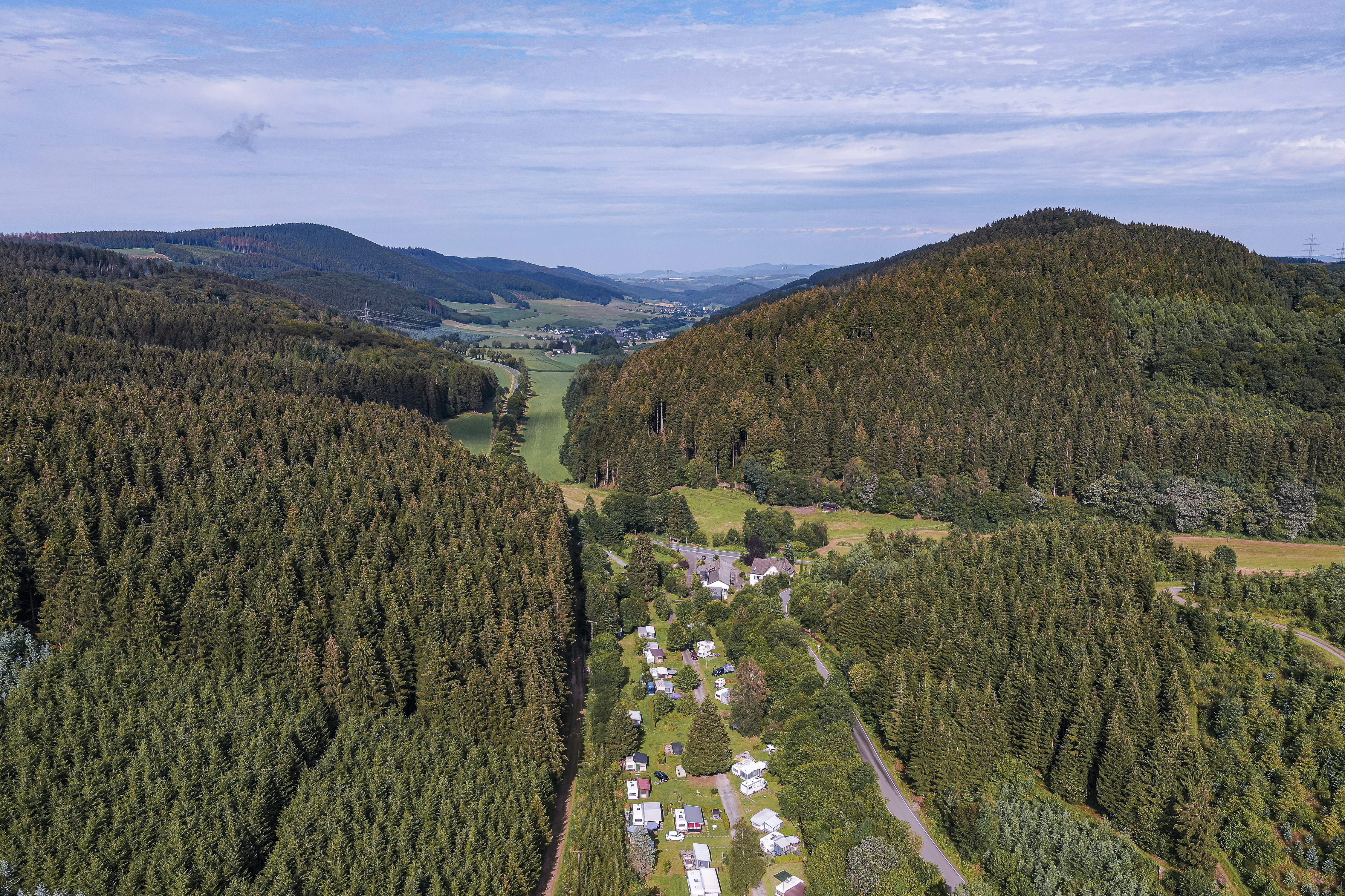Campingplatz im Tal mit kleinen Gebäuden und Stellplätzen, umgeben von dichten Wäldern und Hügeln.