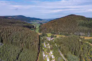 Campingplatz im Tal mit kleinen Gebäuden und Stellplätzen, umgeben von dichten Wäldern und Hügeln.