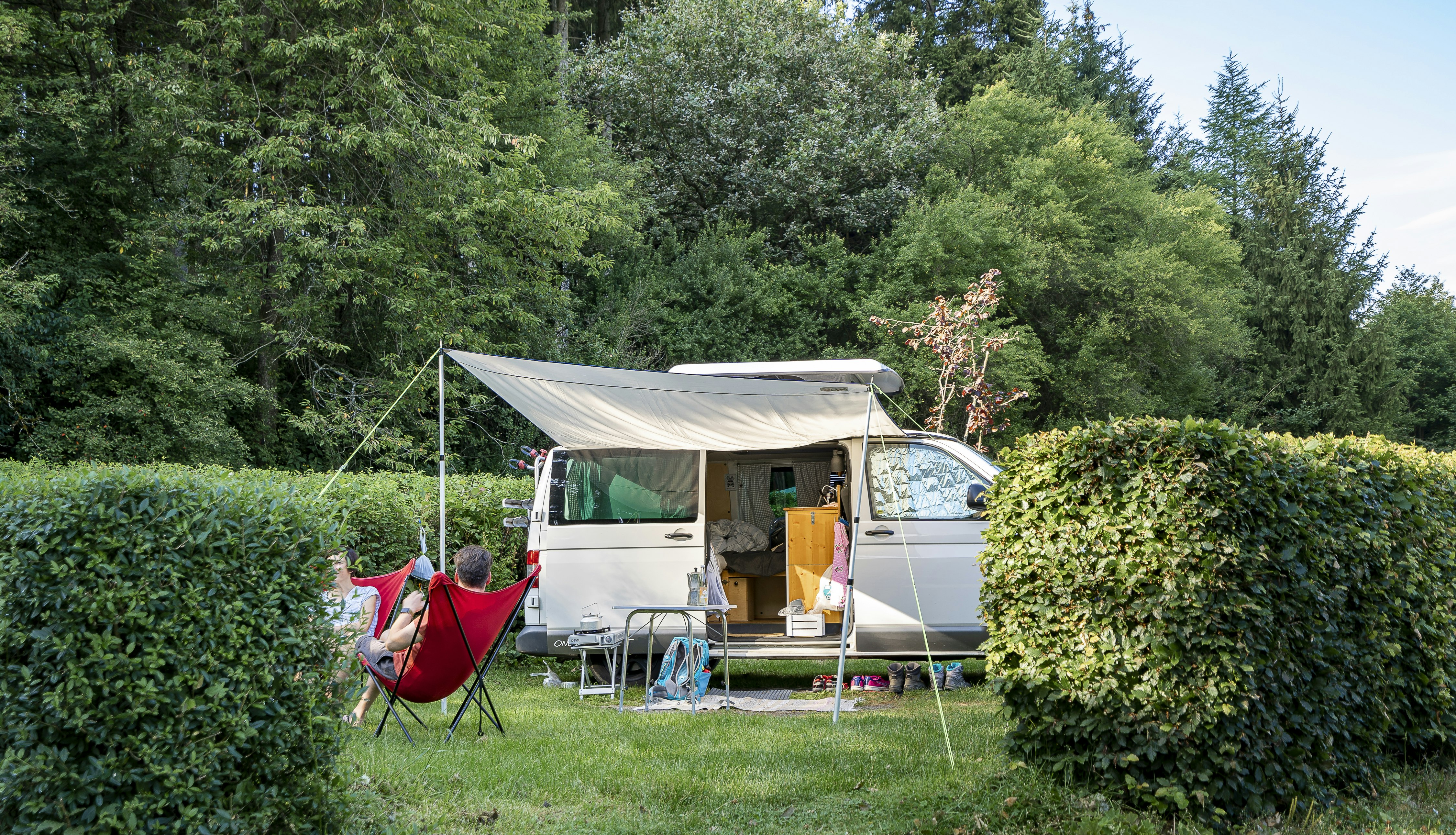 Campingwagen mit geöffnetem Vorzelt, Tisch und Stühlen; daneben eine Hängematte zwischen Büschen und Bäumen.