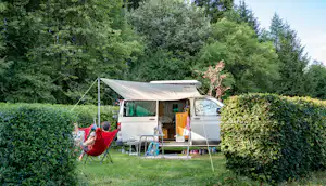 Campingwagen mit geöffnetem Vorzelt, Tisch und Stühlen; daneben eine Hängematte zwischen Büschen und Bäumen.