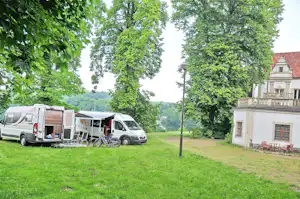 Wohnmobile und ein Campingbus auf einer grünen Wiese, mit Zeltvorbau, Bäumen und einem hellen Gebäude am Rand.