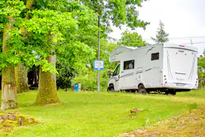 Weißes Wohnmobil auf einem Campingplatz im Grünen neben Bäumen und Rasenfläche, mit Stellplatzschild im Hintergrund.