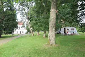 Wohnwagen auf einem Stellplatz im Grünen, umgeben von Bäumen und Rasen, im Hintergrund ein weißes Gebäude.