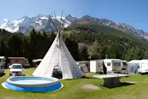 Tipi-Zelt auf dem Campingplatz mit Planschbecken, Wohnwagen und Wohnmobilen im Hintergrund vor Bergpanorama.