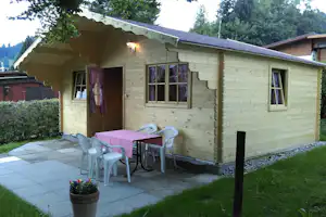 Holzbungalow mit Terrasse, Tisch und Stühlen vor dem Eingang, gepflasterter Bereich und Rasen im Garten, Hecken und Bäume.