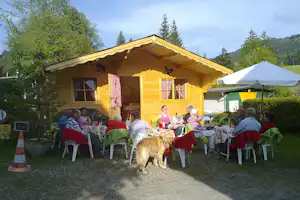 Gelbes Holzhaus mit Terrasse, Gäste sitzen am gedeckten Tisch, daneben ein Hund auf dem Campingplatz im Grünen.