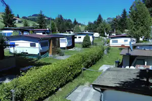Reihen von Wohnwagen und Mobilheimen auf einem Campingplatz mit Hecken, Wegen und Blick auf Bäume und Berge.