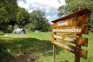 Holz-Wegweiser am Campingplatz mit Aufschriften „Camping“, „Schrottenbaummühle“ und „Insel“; im Hintergrund Zelte und Bäume.