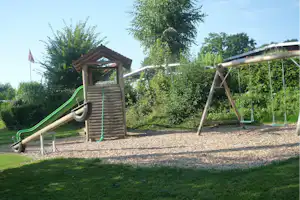 Spielplatz auf dem Campingplatz: grüne Rutsche, Holz-Spielhaus und Schaukeln auf Kiesfläche, umgeben von Bäumen und Sträuchern.