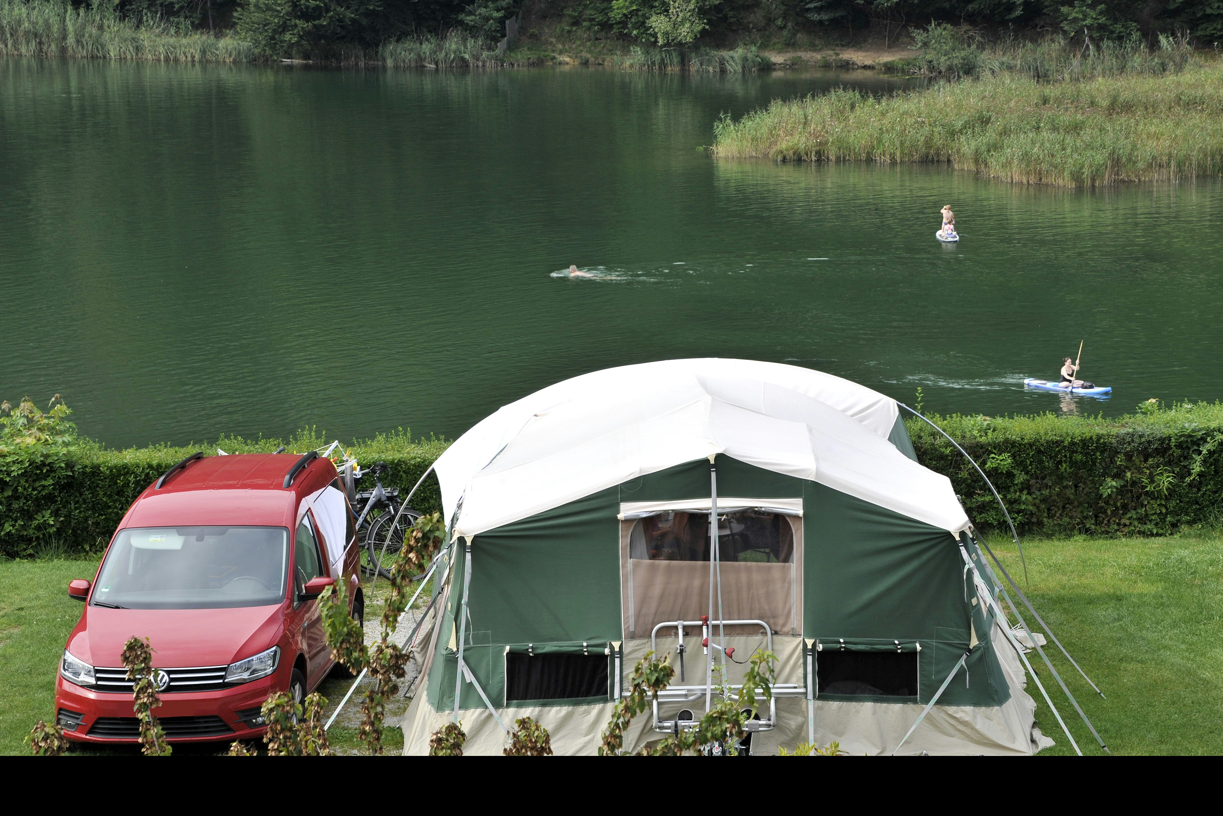 Campingzelt mit Vorhang und Stühlen am Ufer, daneben ein roter Campervan am See, im Hintergrund Bäume.