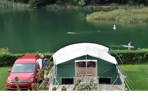 Campingzelt mit Vorhang und Stühlen am Ufer, daneben ein roter Campervan am See, im Hintergrund Bäume.