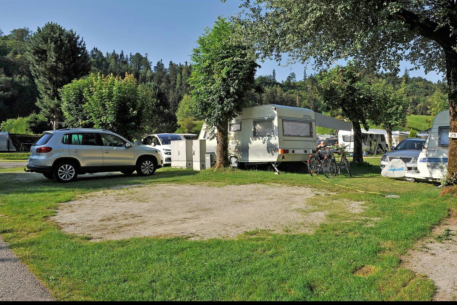 Wohnwagen und Zelte auf einem Campingplatz, mit geparkten Autos, Rasenfläche und Bäumen im Hintergrund.