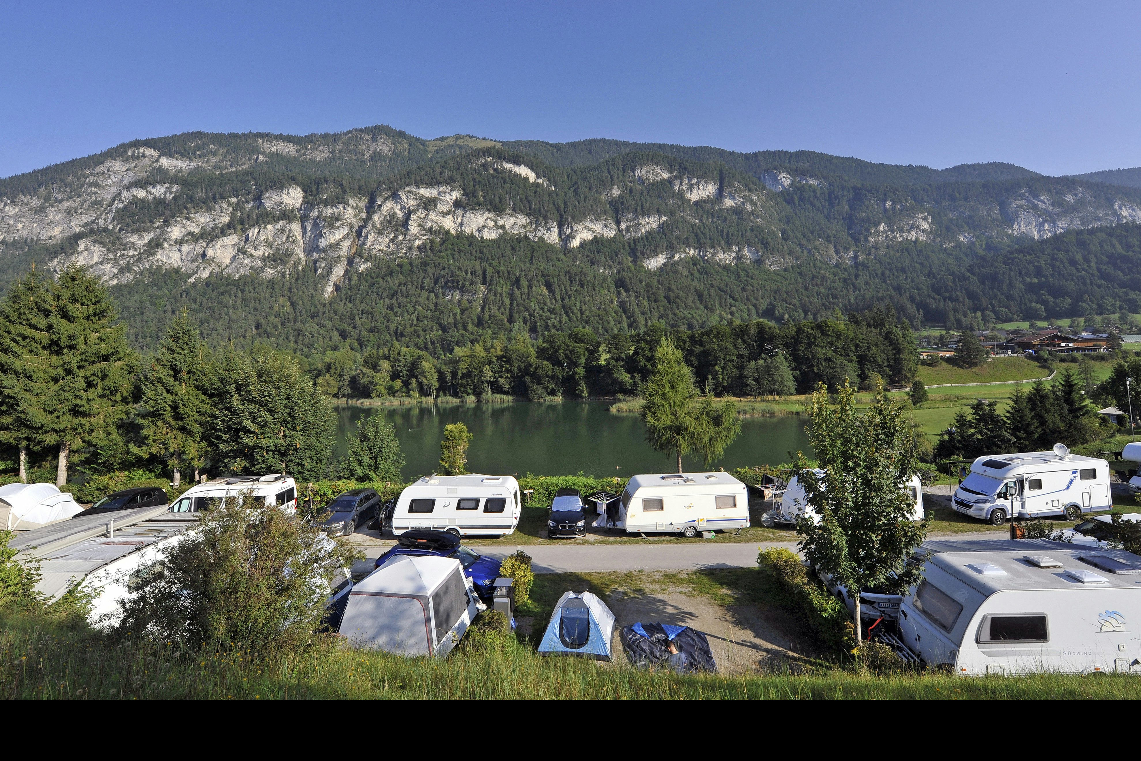 Campingplatz mit Wohnwagen und Zelten auf einem Stellplatz, umgeben von Bäumen und Blick auf bewaldete Berge.