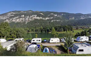 Campingplatz mit Wohnwagen und Zelten auf einem Stellplatz, umgeben von Bäumen und Blick auf bewaldete Berge.