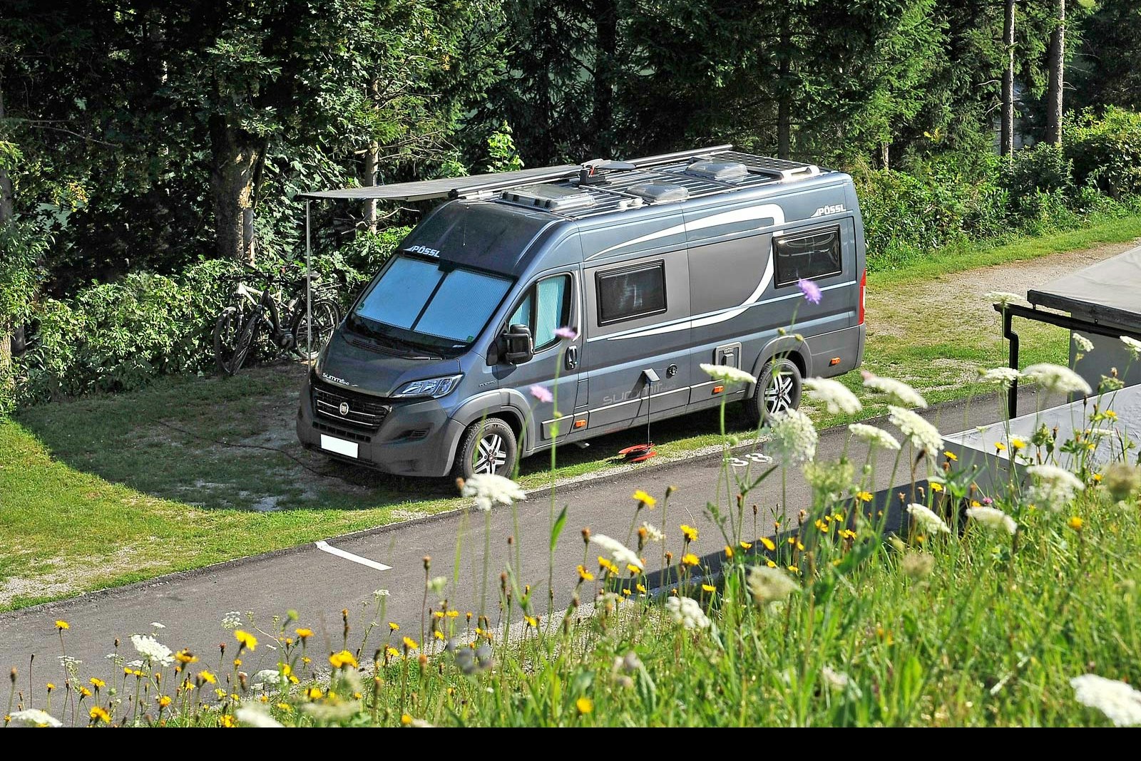Wohnmobil auf einem Stellplatz am Rand eines Rasens, mit Hecke und Bäumen im Hintergrund.