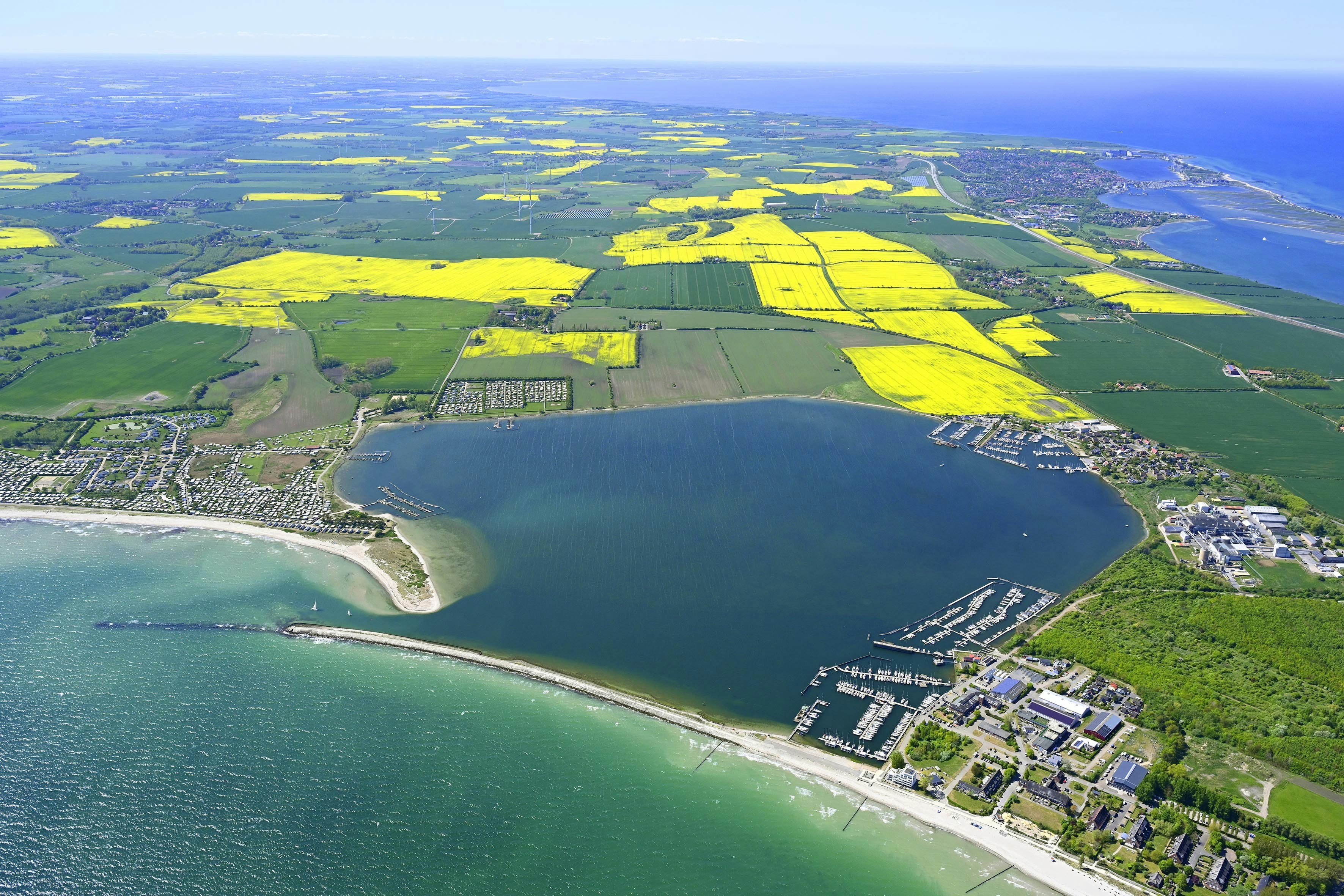 Luftaufnahme eines Sees mit Uferpromenade, Bootsstegen und Wohngebäuden, umgeben von Feldern und Wäldern.