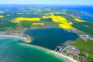 Luftaufnahme eines Sees mit Uferpromenade, Bootsstegen und Wohngebäuden, umgeben von Feldern und Wäldern.