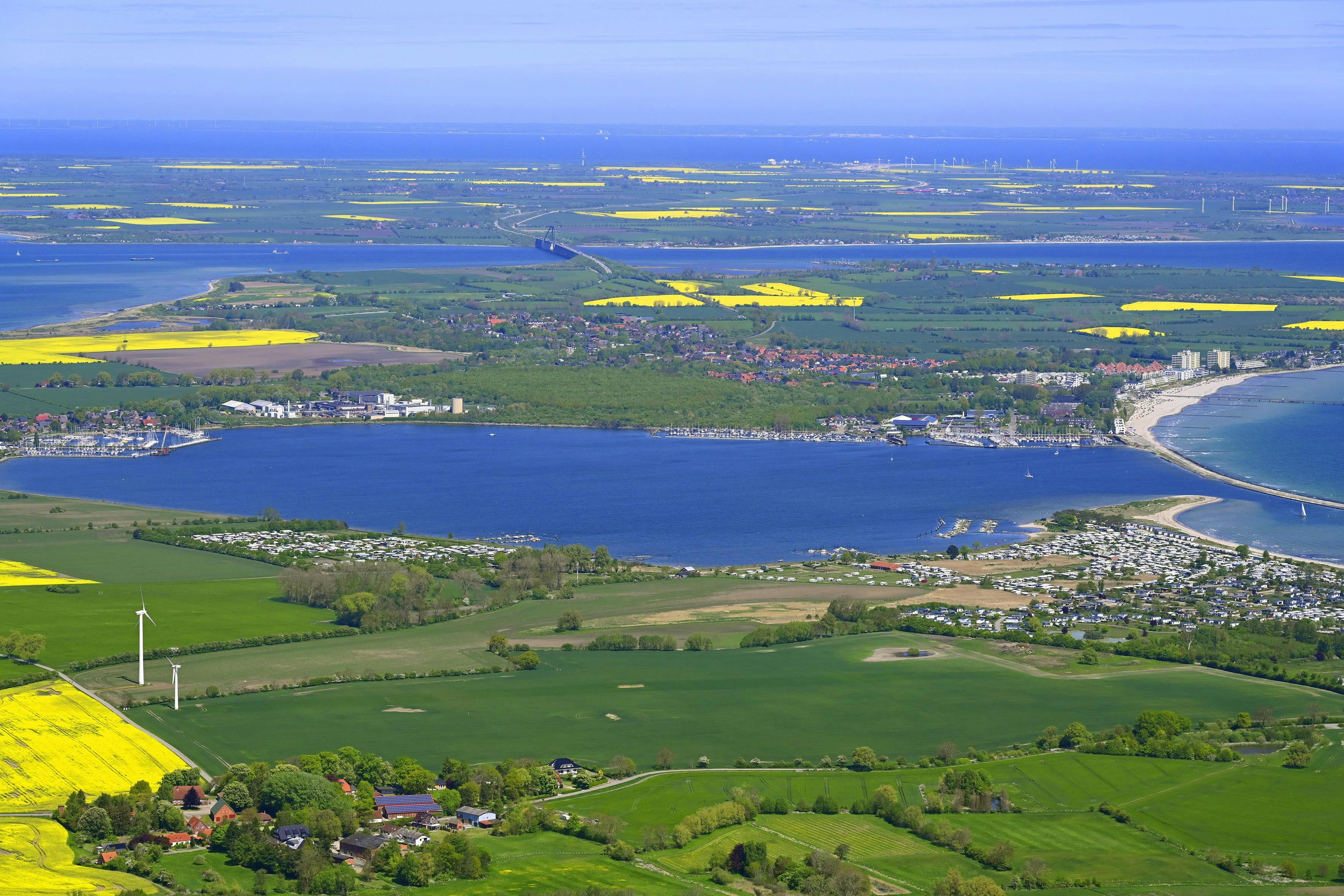 Luftaufnahme einer Küstenlandschaft mit Bucht, Sandstrand, Siedlung und grünen Feldern, dahinter Meer und Himmel.