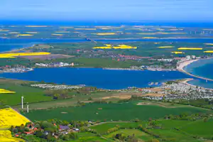 Luftaufnahme einer Küstenlandschaft mit Bucht, Sandstrand, Siedlung und grünen Feldern, dahinter Meer und Himmel.