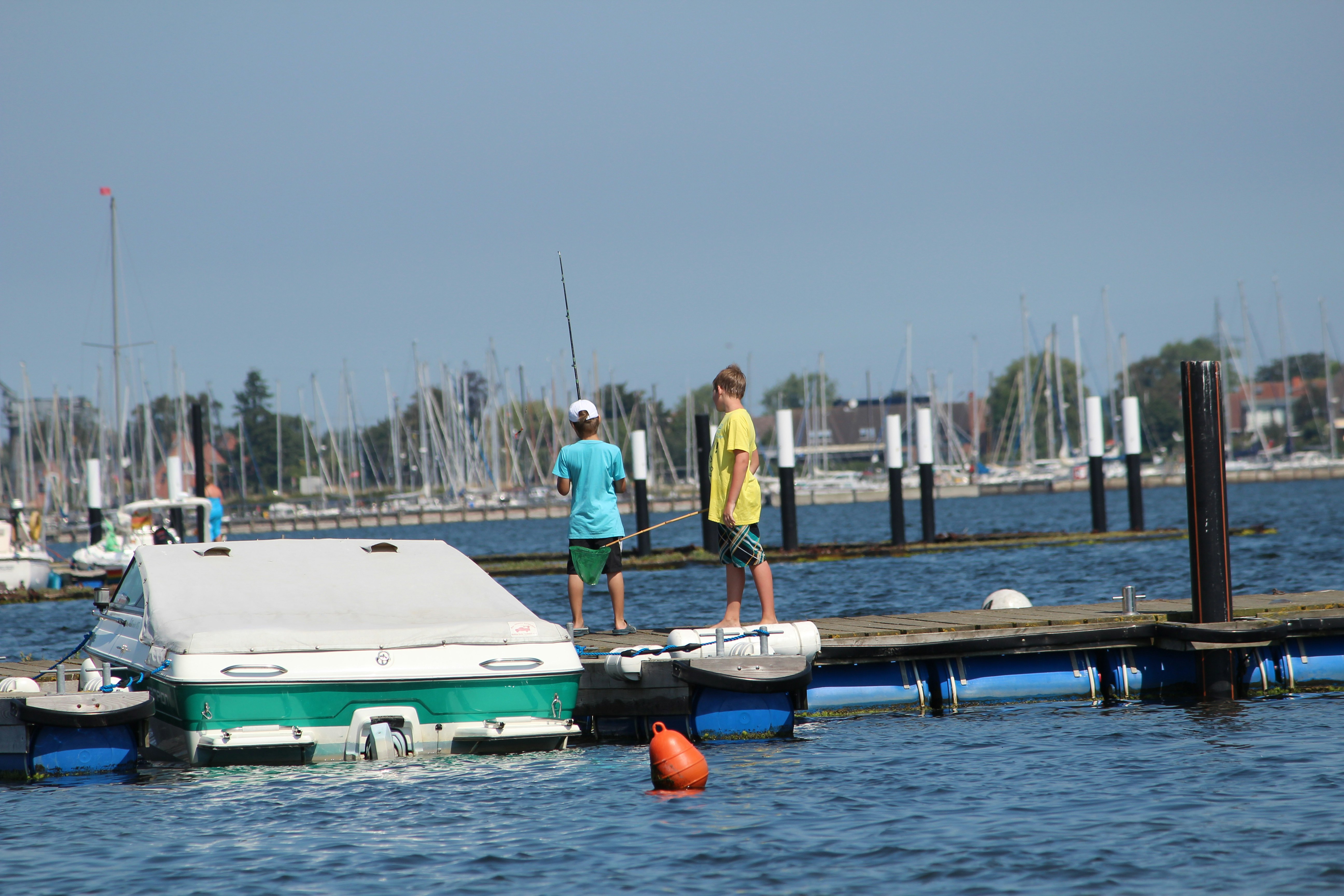 Bootsliegeplatz am Hafen: zwei Personen laufen am Steg entlang, neben einem kleinen Boot und einem orangefarbenen Schwimmkörper.